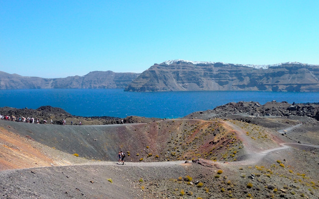 Hikers on volcanic terrain with Santorini caldera and sea in the background.