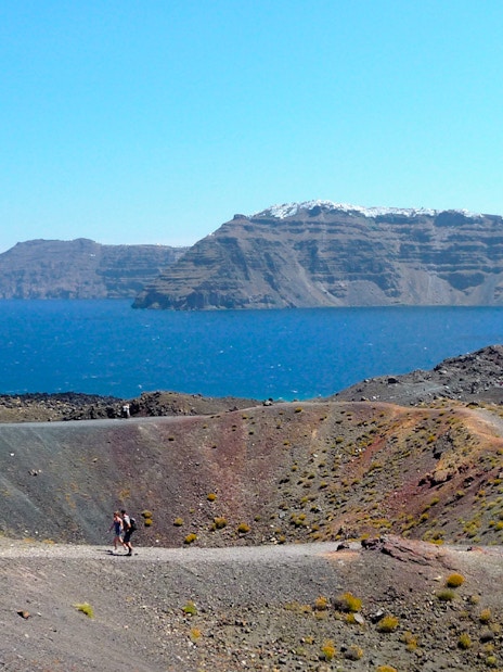 Hikers on volcanic terrain with Santorini caldera and sea in the background.