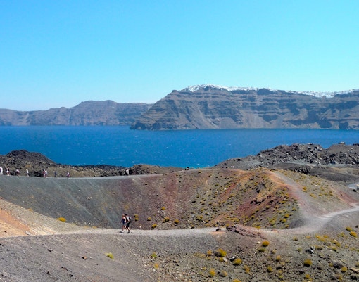 Hikers on volcanic terrain with Santorini caldera and sea in the background.