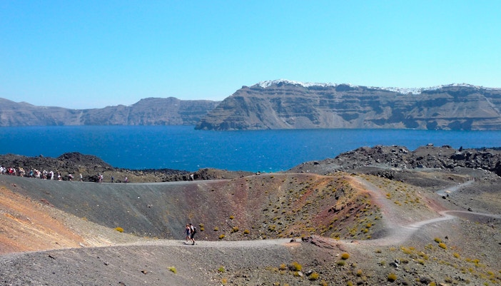 Hikers on volcanic terrain with Santorini caldera and sea in the background.