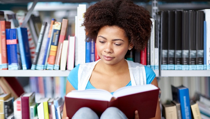 Person reading a book in a library setting, surrounded by shelves of books.