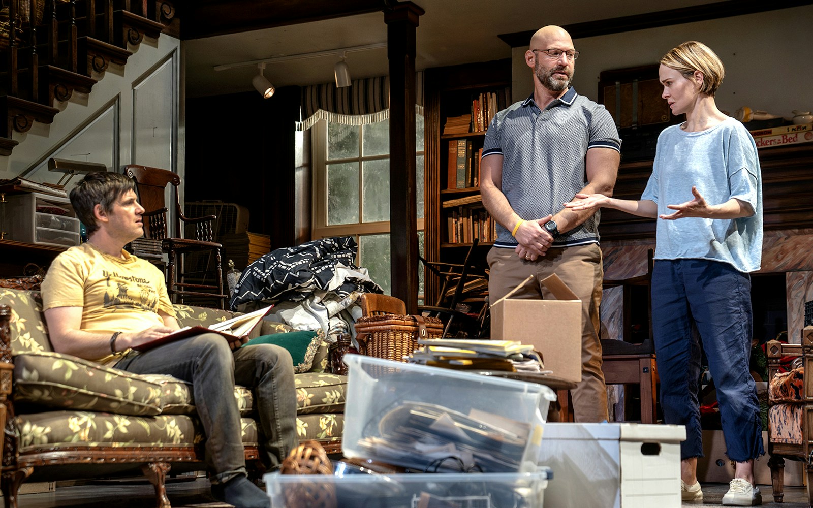 Three people in a cluttered living room, engaged in conversation, surrounded by boxes and furniture.