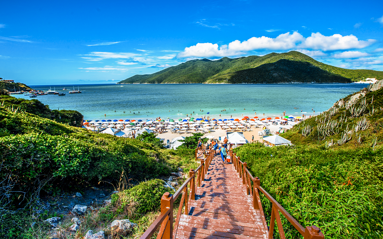 Wooden staircase leading to Pontal do Atalaia Beach, Arraial do Cabo, with colorful umbrellas and ocean view.