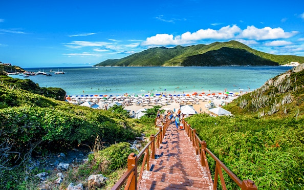 Wooden staircase leading to Pontal do Atalaia Beach, Arraial do Cabo, with colorful umbrellas and ocean view.
