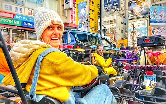 Woman in a white beanie and yellow hoodie driving a go-kart on a busy Tokyo street.