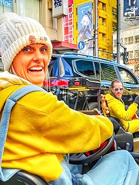 Woman in a white beanie and yellow hoodie driving a go-kart on a busy Tokyo street.