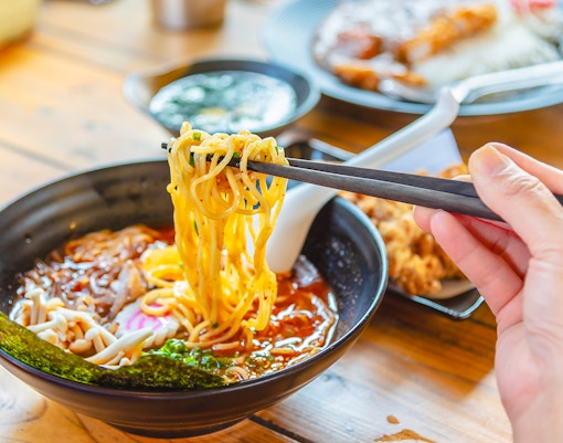 Chopsticks lifting noodles from a bowl of ramen with toppings on a wooden table.