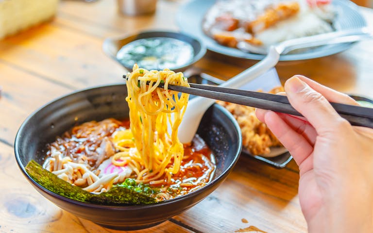 Chopsticks lifting noodles from a bowl of ramen with toppings on a wooden table.