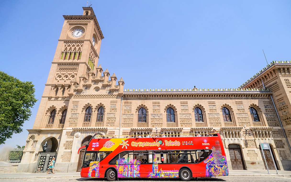 Red Toledo sightseeing bus in front of historic building with clock tower.