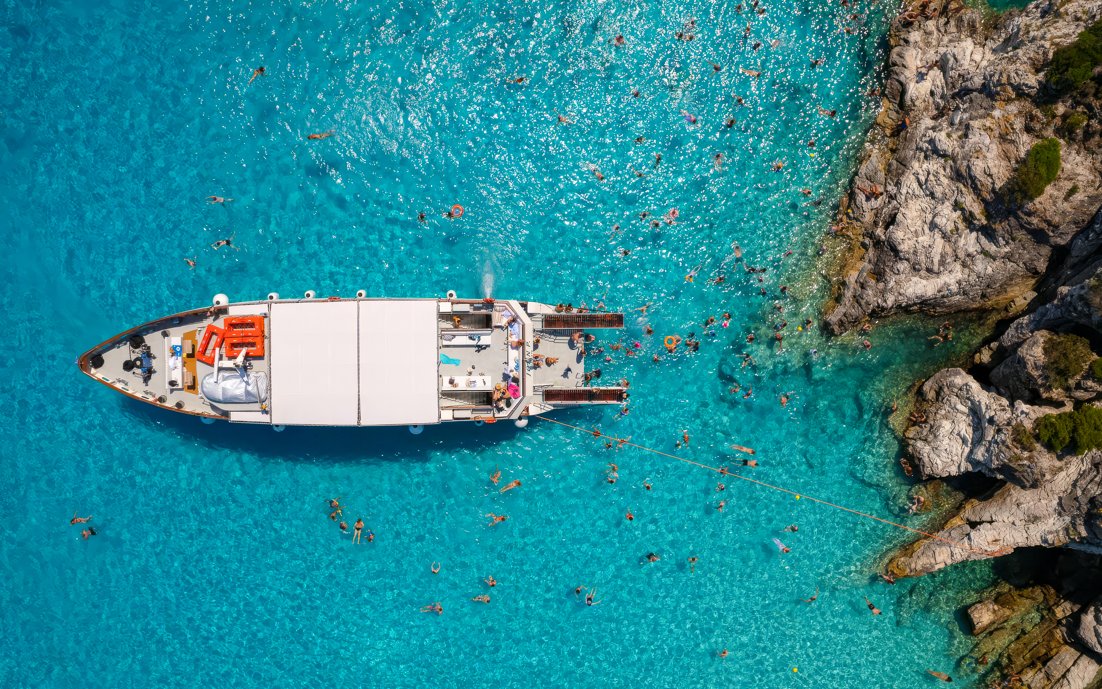 Boats anchored near rocky shore in Paxos and Antipaxos with swimmers in clear blue water.
