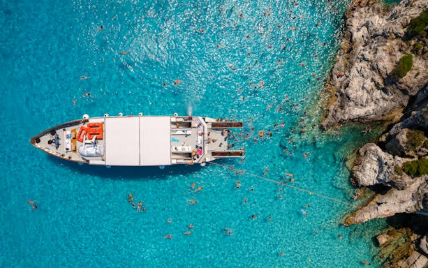 Boats anchored near rocky shore in Paxos and Antipaxos with swimmers in clear blue water.