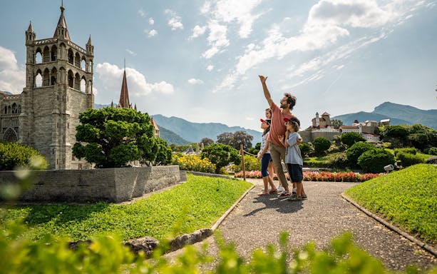Family exploring miniature landmarks at Swissminiatur Museum with mountains in the background.
