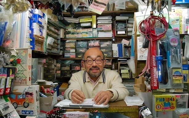 Stationery shop owner at Akihabara surrounded by various office supplies.