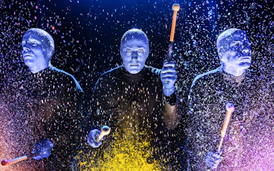 Blue Man Group performers playing drums with colorful splashes on stage.