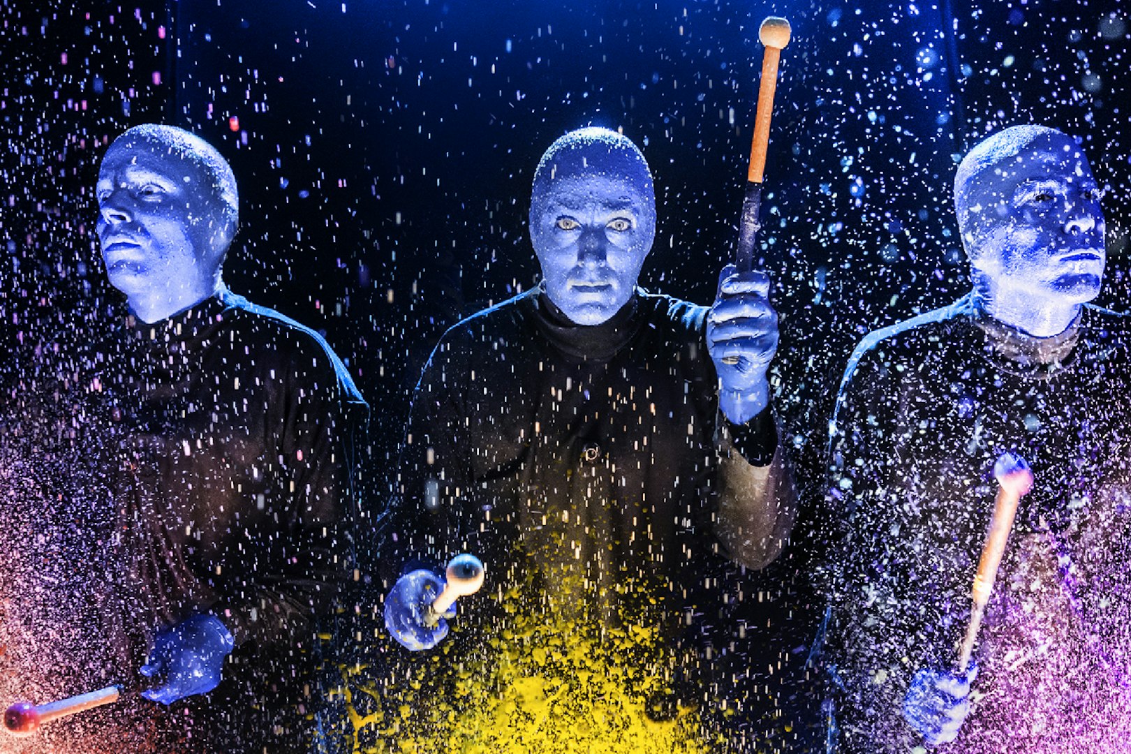 Blue Man Group performers playing drums with colorful splashes on stage.