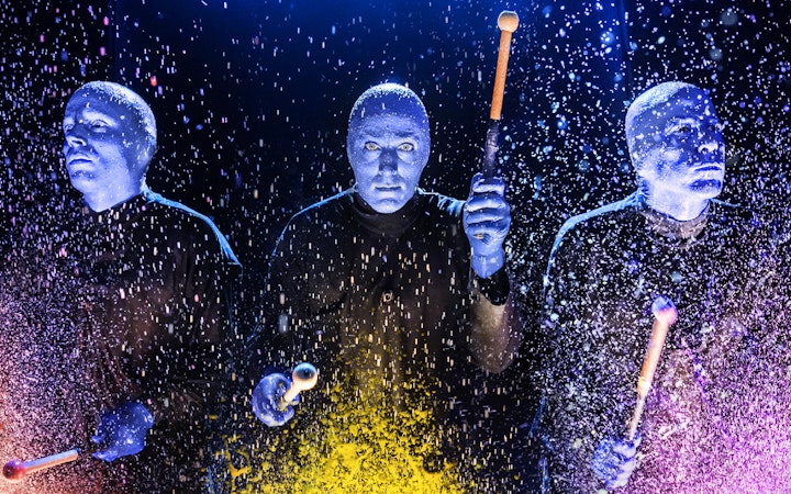 Blue Man Group performers playing drums with colorful splashes on stage.