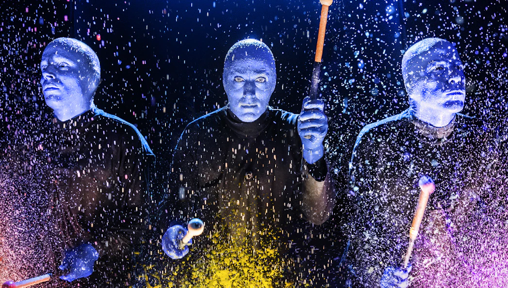 Blue Man Group performers playing drums with colorful splashes on stage.