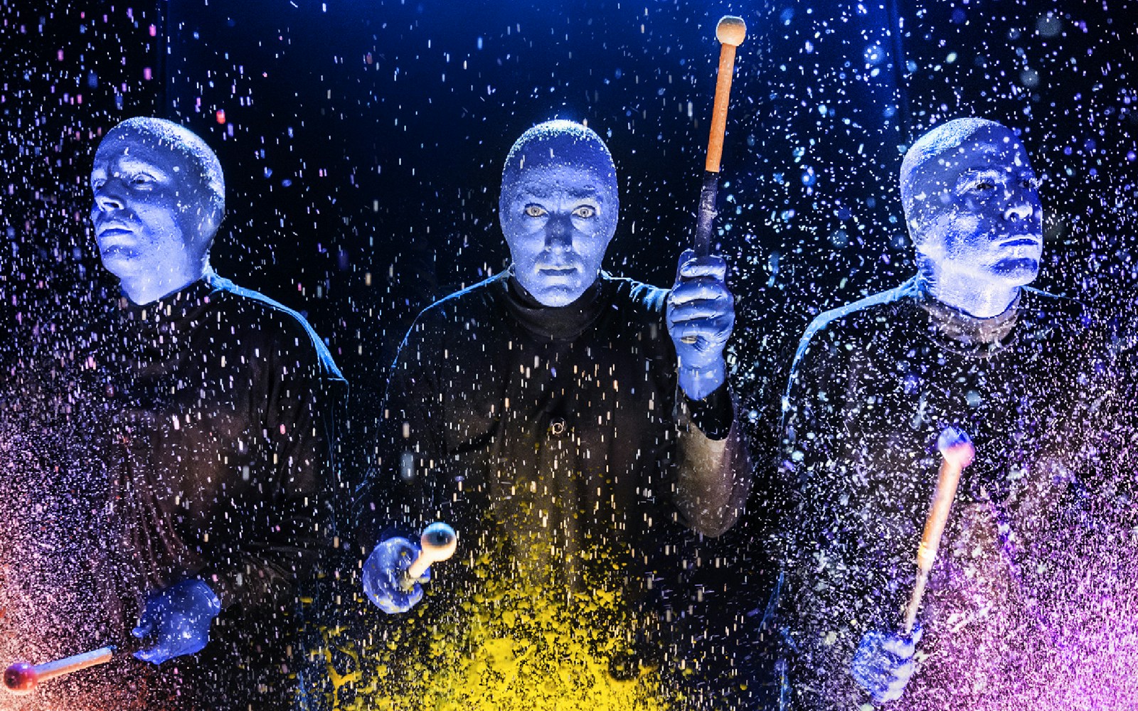 Blue Man Group performers playing drums with colorful splashes on stage.