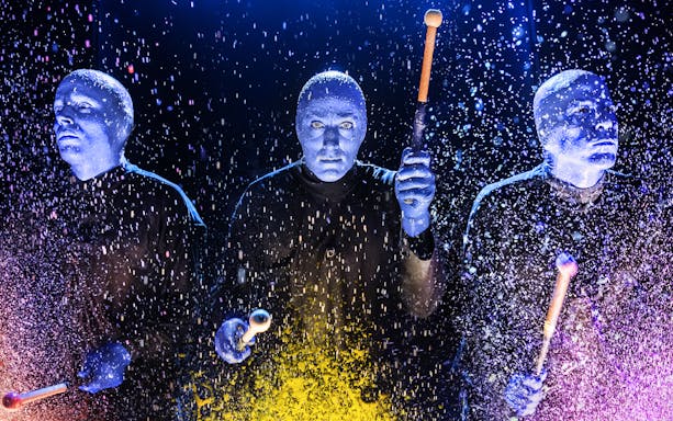 Blue Man Group performers playing drums with colorful splashes on stage.