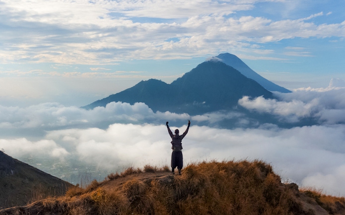 Man raising arms while viewing Mount Batur during sunrise trek in Bali.