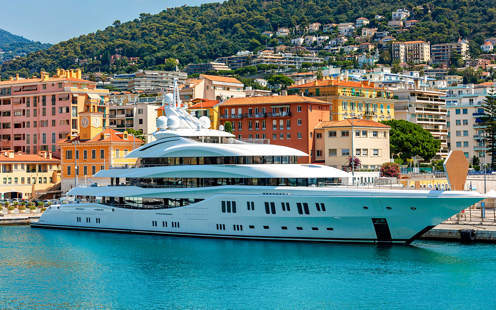 Luxury yacht docked at Port de Nice with colorful buildings and hills in the background.