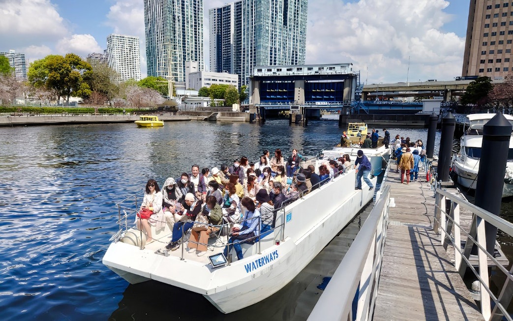 Passengers boarding a cruise boat on the Meguro River in Tokyo with cityscape in the background.