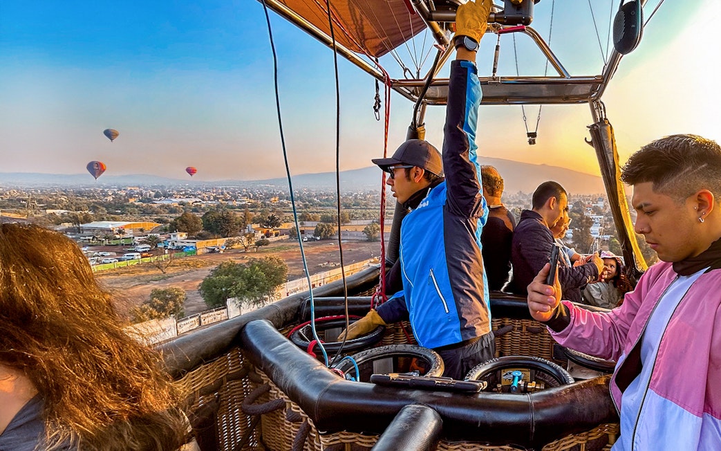 Pilot and passengers in a hot air balloon over Teotihuacan with distant balloons in the sky.