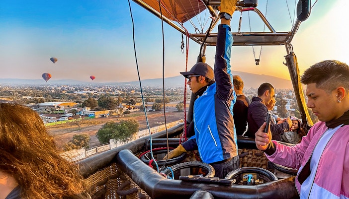 Pilot and passengers in a hot air balloon over Teotihuacan with distant balloons in the sky.