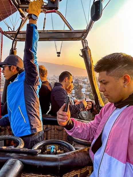 Pilot and passengers in a hot air balloon over Teotihuacan with distant balloons in the sky.