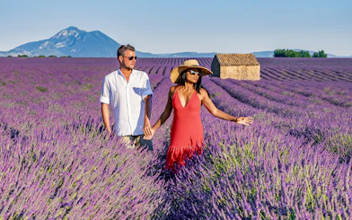 Couple walking through a lavender field in Provence with a mountain and stone house in the background.