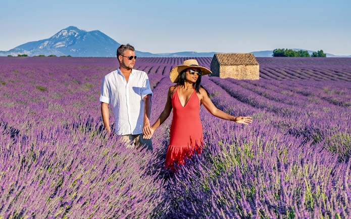 Couple walking through a lavender field in Provence with a mountain and stone house in the background.