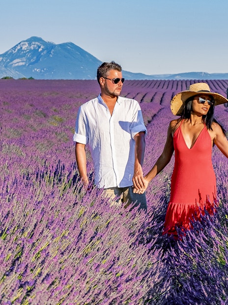 Couple walking through a lavender field in Provence with a mountain and stone house in the background.