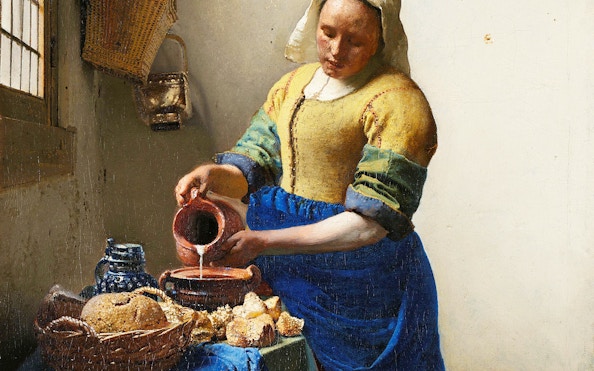 Milkmaid pouring milk into a bowl beside bread and pottery in a rustic kitchen.