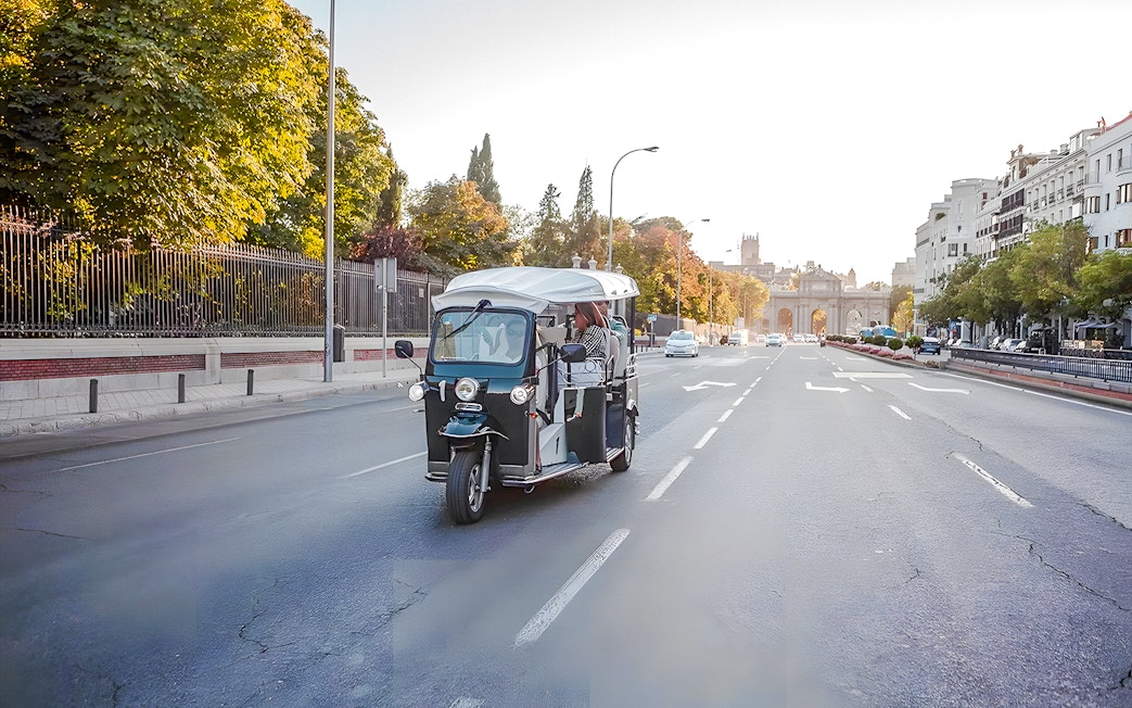 Electric tuk-tuk on a Madrid street with Puerta de Alcalá in the background.