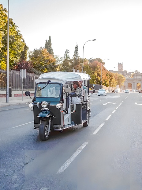 Electric tuk-tuk on a Madrid street with Puerta de Alcalá in the background.
