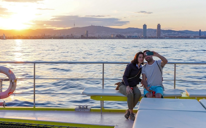Couple taking a selfie on a catamaran cruise with Barcelona skyline at sunset.