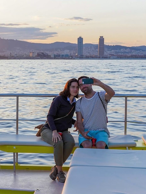 Couple taking a selfie on a catamaran cruise with Barcelona skyline at sunset.