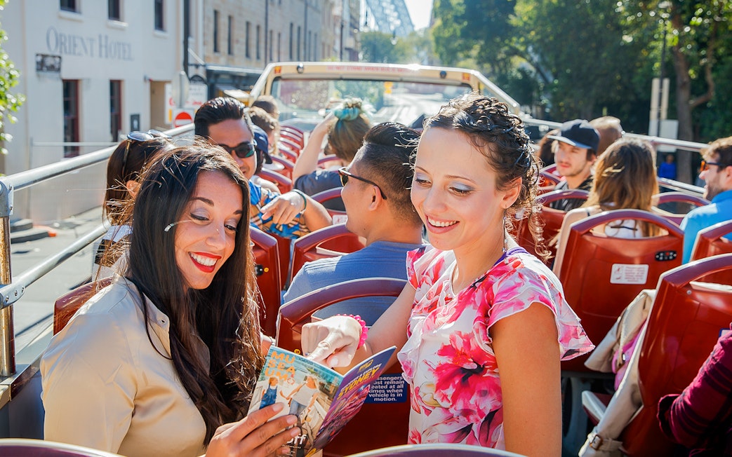 Two women reading a map on an open-top bus tour in a city.