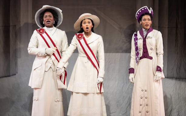 Three performers in period costumes on stage during a scene from "Suffs" musical.
