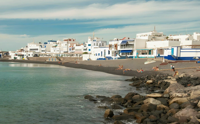 Dark-sand beach at Playa del Muellito, El Cotillo, Fuerteventura, with coastal village buildings.