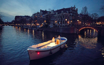 Cloud Boat Cruise on Amsterdam canal at dusk with lit bridge and historic buildings.