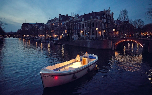 Cloud Boat Cruise on Amsterdam canal at dusk with lit bridge and historic buildings.