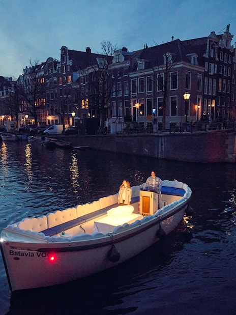 Cloud Boat Cruise on Amsterdam canal at dusk with lit bridge and historic buildings.