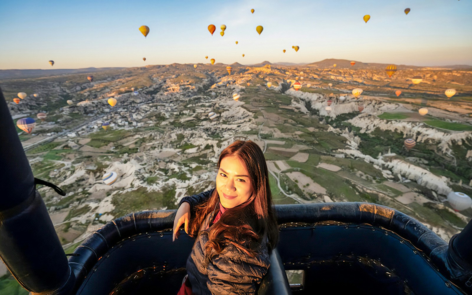 Woman in hot air balloon over Cappadocia landscape with multiple balloons in the sky.