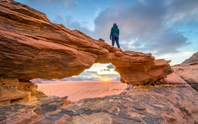 Person standing on rock arch at sunset in Wadi Rum desert, Jordan.
