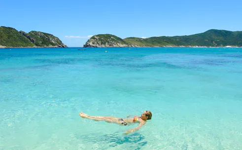 Person floating in clear turquoise water at Arraial do Cabo, Brazil, with green hills in the background.