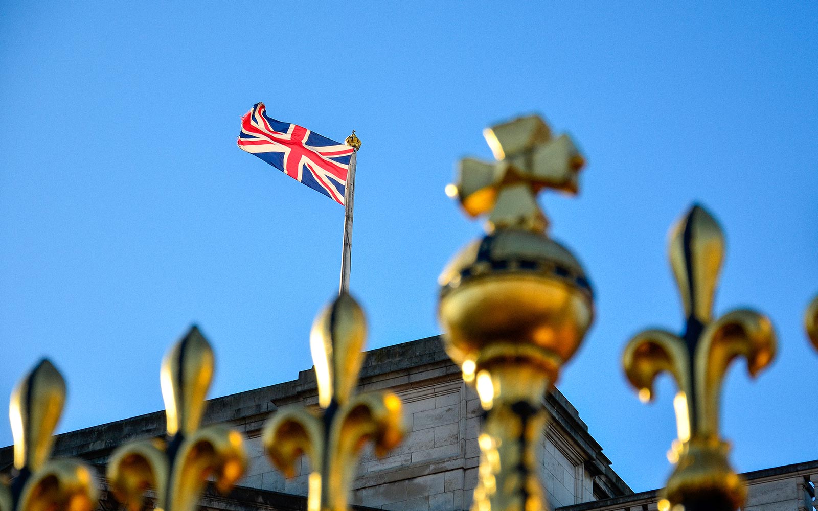 Union Jack flag flying above Buckingham Palace with golden fence in foreground.