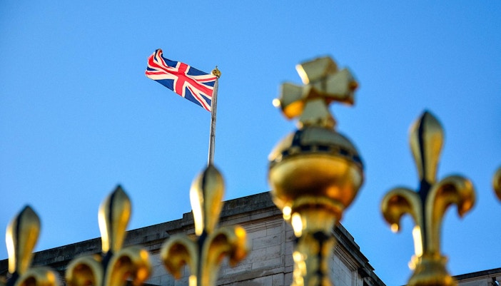 Union Jack flag flying above Buckingham Palace with golden fence in foreground.