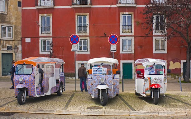 Three electric tuk tuks parked in front of a red building in Lisbon for a guided sightseeing tour.
