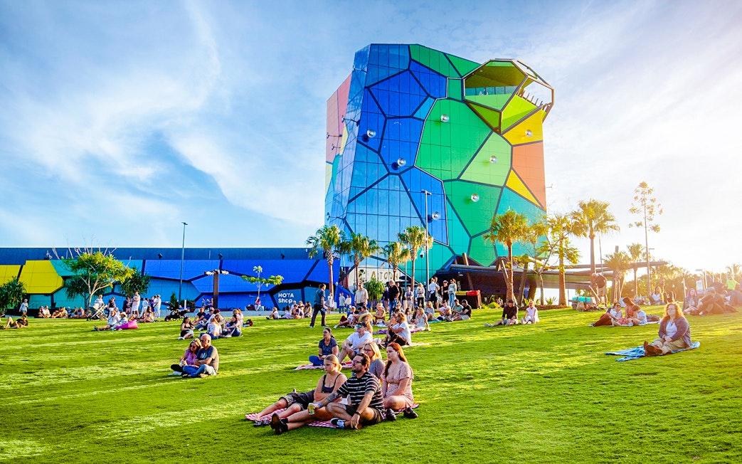 People relaxing on the lawn at Home of the Arts, Gold Coast, with colorful building in background.